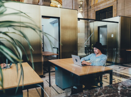 A man in a beanie working on a laptop at a desk in an open office space